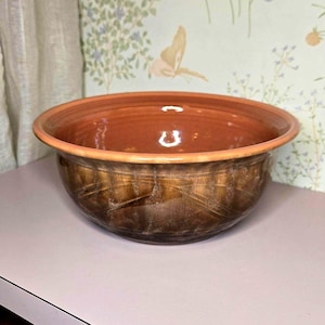 May include: A round ceramic bowl with a smooth, glossy terracotta-colored interior and a wide, rounded rim. The exterior features a dark, mottled brown glaze with subtle, textured diagonal striations that wrap around the curved body.