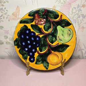 May include: A hand-painted ceramic plate with a vibrant yellow background. The plate features a design of grapes, a pear, a strawberry, and a peach, all in vivid colors. The plate is displayed on a decorative gold-colored stand.