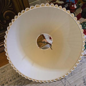 May include: Cream-coloured lampshade with a scalloped edge. The lampshade has a wide, conical shape and a central opening for the light fixture. The edge is trimmed with a decorative, wavy pattern. The lampshade is made of a soft, opaque material.