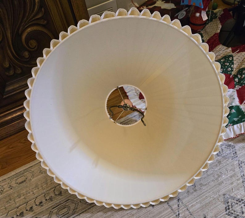 May include: Cream-coloured lampshade with a scalloped edge. The lampshade has a wide, conical shape and a central opening for the light fixture. The edge is trimmed with a decorative, wavy pattern. The lampshade is made of a soft, opaque material.