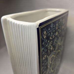 May include: A rectangular ceramic vase with a white interior and a ribbed white side edge. The front panel features a dark blue background with a detailed, intricate floral and geometric pattern in shades of light blue, cream, and gold-toned accents.