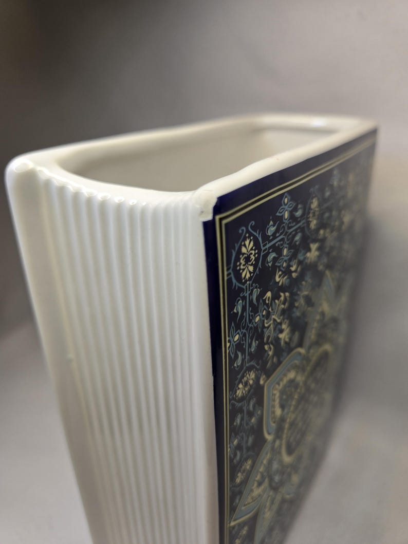May include: A rectangular ceramic vase with a white interior and a ribbed white side edge. The front panel features a dark blue background with a detailed, intricate floral and geometric pattern in shades of light blue, cream, and gold-toned accents.