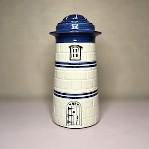 May include: A ceramic lighthouse-shaped container with a blue top and accents. The body is white with brick-like detailing and blue stripes. Features a small window and door design. A decorative nautical-themed item.
