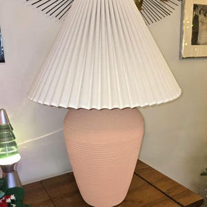 May include: A peach-coloured ceramic table lamp with a cream-coloured pleated lampshade. The lamp base has a textured, ribbed design. The lamp is on a wooden surface.