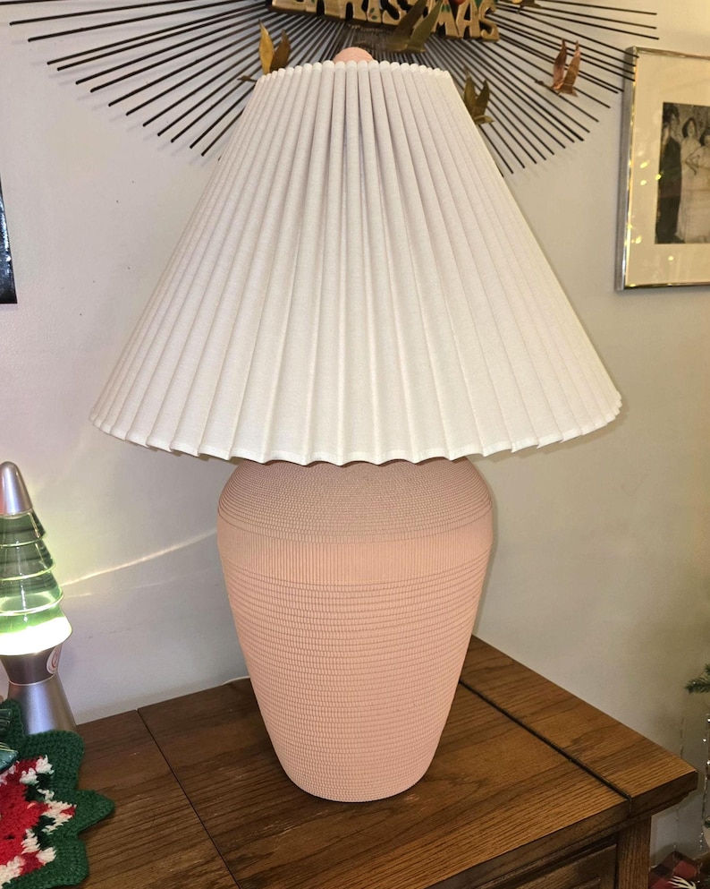 May include: A peach-coloured ceramic table lamp with a cream-coloured pleated lampshade. The lamp base has a textured, ribbed design. The lamp is on a wooden surface.