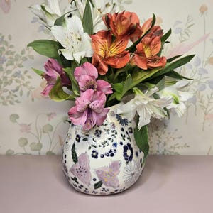 May include: A floral arrangement in a decorative vase. The bouquet features white, orange, and pink flowers with green leaves. The vase is white with a floral pattern in blue and purple.
