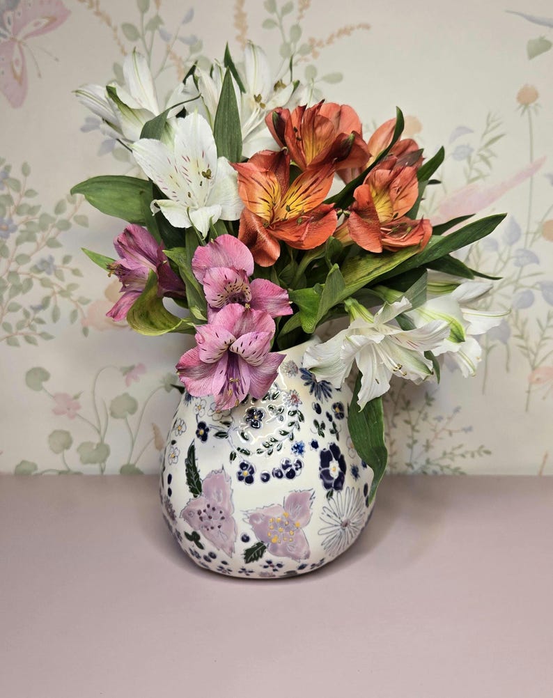 May include: A floral arrangement in a decorative vase. The bouquet features white, orange, and pink flowers with green leaves. The vase is white with a floral pattern in blue and purple.