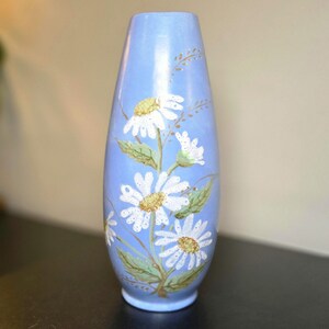 May include: A light blue vase with a tapered cylindrical shape, featuring a hand-painted design of white daisy flowers with yellow centers, green leaves, and thin brown stems extending vertically along the front surface of the ceramic body.