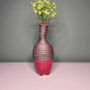 May include: A decorative ceramic vase with a red base and a brown, ridged body. The vase holds a bouquet of small, light green flowers. The vase is on a pink surface against a gray background.