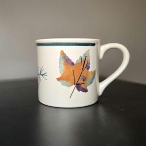 May include: White ceramic mug with a teal blue stripe near the rim and a leaf design in orange, purple, and teal. The mug has a curved handle and a simple, elegant design.