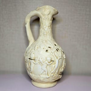 May include: Off-white ceramic pitcher with a handle and a detailed relief design. The relief depicts figures, trees, and floral patterns. The pitcher has a rounded base and a spout with a decorative edge.