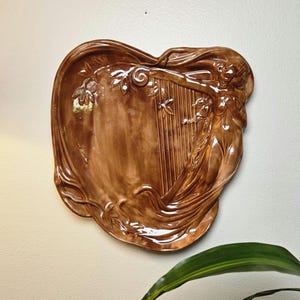 May include: A glossy brown ceramic wall plaque featuring a relief sculpture of a figure playing a harp. The design includes flowing drapery and leaf motifs integrated into the irregular, organic shape of the piece, creating a textured surface.