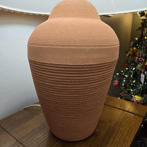 May include: A terracotta-coloured table lamp with a textured, ribbed design. The lamp has a rounded top and a wide base, topped with a white lampshade. The lamp sits on a wooden table.