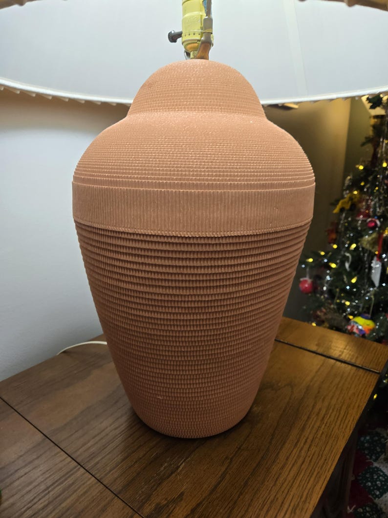 May include: A terracotta-coloured table lamp with a textured, ribbed design. The lamp has a rounded top and a wide base, topped with a white lampshade. The lamp sits on a wooden table.