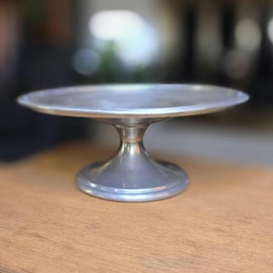 May include: A silver-colored, vintage cake stand with a round top and a pedestal base. The stand is made of metal and sits on a wooden surface. The design is simple and elegant, suitable for displaying baked goods.