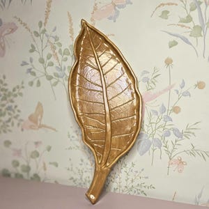 May include: A gold-colored leaf-shaped decorative item. The leaf has detailed veining and a textured surface. The item is mounted on a wall with a floral and butterfly wallpaper background. The leaf is a decorative accent.
