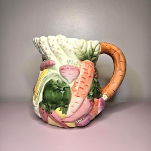 May include: A ceramic pitcher with a handle, decorated with colorful vegetables. The pitcher features a carrot-shaped handle and is adorned with images of asparagus, corn, carrots, and other vegetables. The design is detailed and vibrant, with a glossy finish.