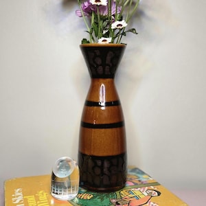May include: A brown and black ceramic vase with a flared top, holding purple and white flowers. The vase sits on a stack of books, with a clear glass object in front. The book has the text "With Skies and Wings".