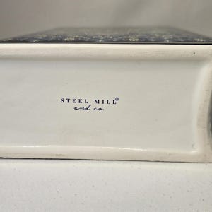 May include: A white ceramic object shaped like a book with a dark blue patterned top surface. The side of the object features the text STEEL MILL and co. printed in black. The ceramic has a smooth finish and a curved spine-like edge.