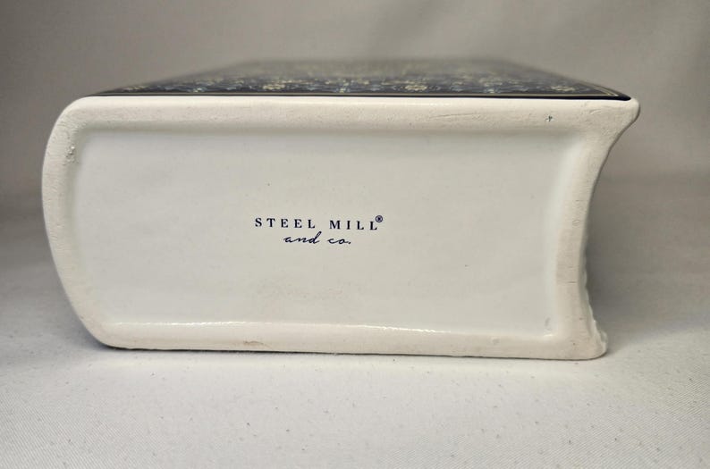 May include: A white ceramic object shaped like a book with a dark blue patterned top surface. The side of the object features the text STEEL MILL and co. printed in black. The ceramic has a smooth finish and a curved spine-like edge.