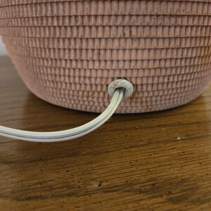 May include: Close-up of a pink, textured lamp base with a white electrical cord extending from the bottom. The lamp base has a basket-weave design. The cord is plugged into the base.