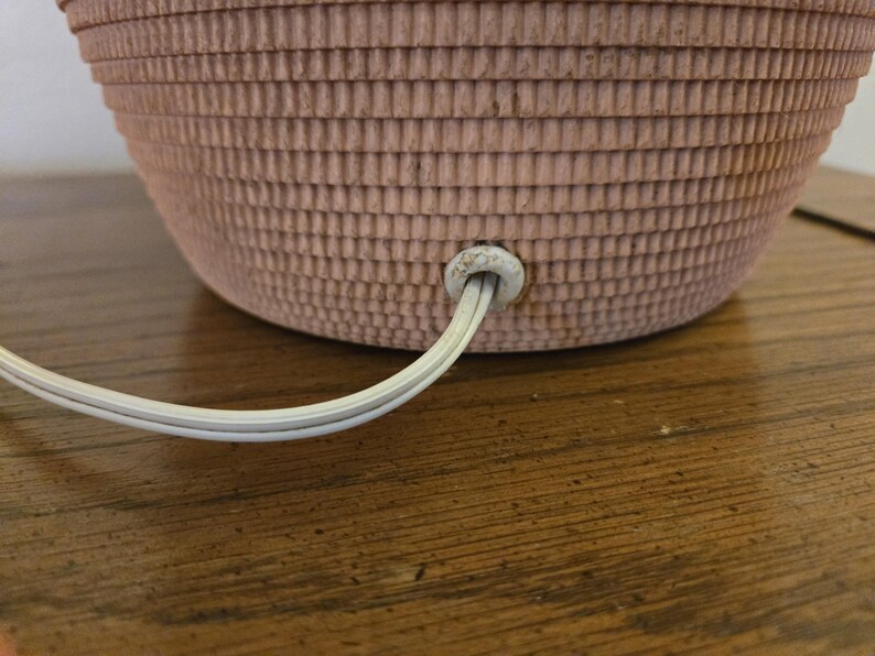 May include: Close-up of a pink, textured lamp base with a white electrical cord extending from the bottom. The lamp base has a basket-weave design. The cord is plugged into the base.