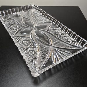 May include: A rectangular clear glass serving tray featuring an intricate pressed pattern of a central floral motif surrounded by geometric starbursts and cross-hatched textures. The tray has a scalloped, serrated edge and a raised rim.