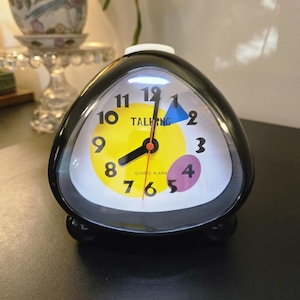 May include: A black, triangular-shaped quartz alarm clock with a white face. The dial features black numbers, a yellow circular center, a blue triangle, and a purple oval. The text "TALKING" and "QUARTZ ALARM" are printed in black on the clock face.