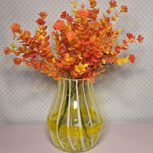 May include: A clear glass vase with yellow and white stripes holds a vibrant arrangement of orange and yellow artificial flowers. The flowers have small, rounded leaves and are arranged to create a full, textured display. The vase sits on a pink surface.