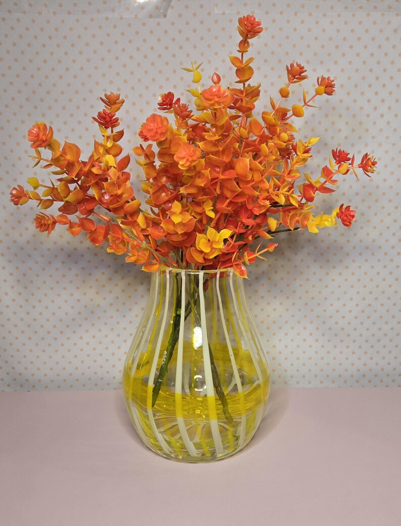 May include: A clear glass vase with yellow and white stripes holds a vibrant arrangement of orange and yellow artificial flowers. The flowers have small, rounded leaves and are arranged to create a full, textured display. The vase sits on a pink surface.