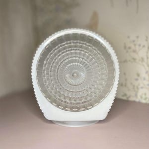 May include: A white, circular light fixture with a clear, textured glass shade. The glass has a radial pattern emanating from the center. The fixture has a scalloped edge and a rounded base, suitable for a ceiling or wall mount.