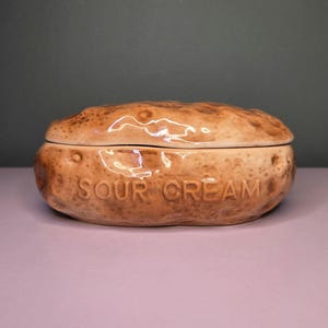 May include: A ceramic sour cream dish with a lid. The dish is shaped like a potato and is light brown with darker brown speckles. The words "SOUR CREAM" are embossed on the front of the dish.