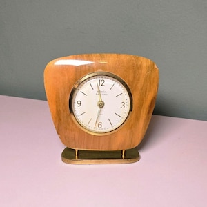 May include: A vintage wooden clock with a square face and rounded edges. The clock face is white with black numerals and gold hands. The clock sits on a gold-colored base and has a light brown wooden frame.