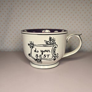 May include: White ceramic mug with a purple interior and a black rim. The mug features the phrase "do your BEST" in black script, framed by floral illustrations. The handle is curved and the mug sits on a small base.