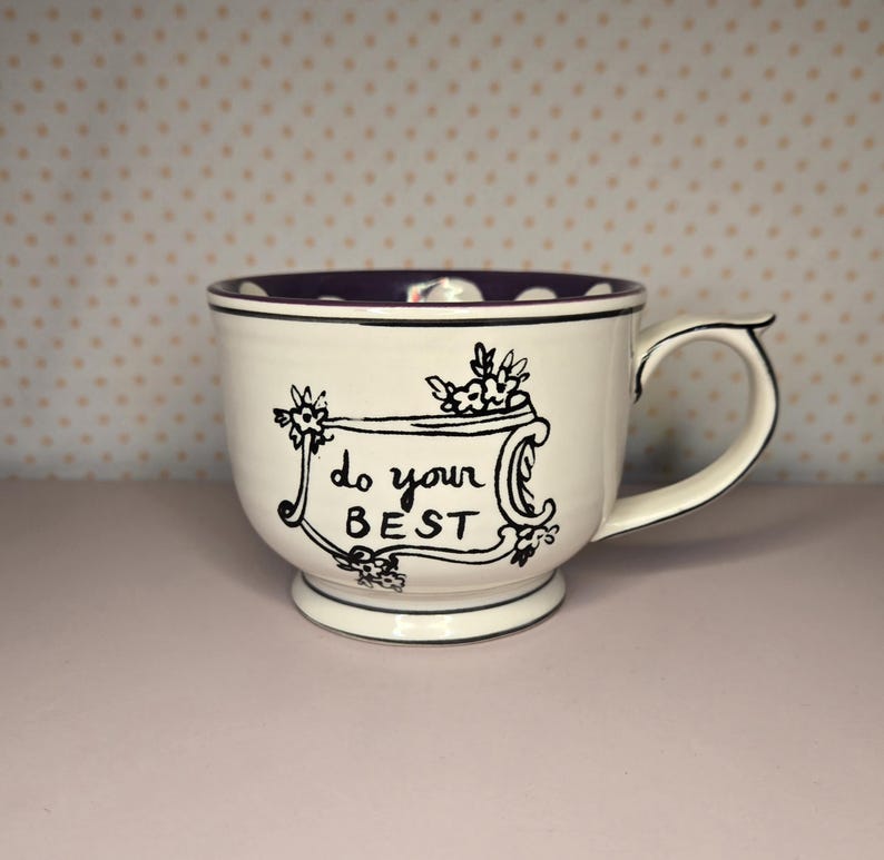 May include: White ceramic mug with a purple interior and a black rim. The mug features the phrase "do your BEST" in black script, framed by floral illustrations. The handle is curved and the mug sits on a small base.