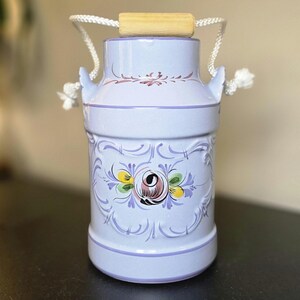 May include: A light purple ceramic milk can featuring a hand-painted floral motif in shades of pink, yellow, and green. The vessel includes a white rope handle with a wooden grip attached to two side lugs, and a decorative purple scrollwork border.