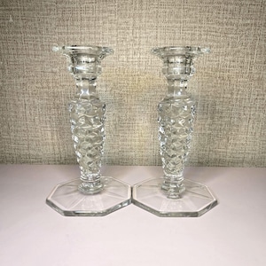 Puede incluir: Dos candelabros de cristal transparente. Cada uno tiene una base hexagonal, un tallo facetado y una parte superior acampanada. Los candelabros están hechos de cristal transparente y se exhiben sobre una superficie blanca contra un fondo texturizado.