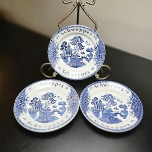 May include: Three white ceramic plates featuring a blue willow pattern and the word Schweppes printed in black text along the rim. One plate is displayed on a silver metal stand, while two identical plates are positioned in front of the stand.