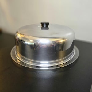 May include: A round, silver-colored metal food cover with a smooth, reflective surface and a small, black, cylindrical knob handle centered on the top. The dome-shaped cover sits securely on a matching circular metal base plate with a flat rim.