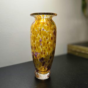 May include: A hand-blown glass vase featuring a speckled pattern of amber, gold, and deep purple spots against a clear base. The vessel has a flared rim, a tapered cylindrical body, and a thick, clear glass circular base for stability.
