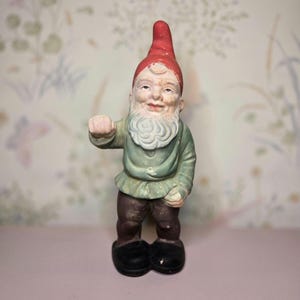 May include: A ceramic garden gnome figurine. The gnome wears a red pointed hat, a green shirt, brown pants, and black shoes. The gnome has a white beard and is making a fist with one hand. The background is a floral wallpaper.