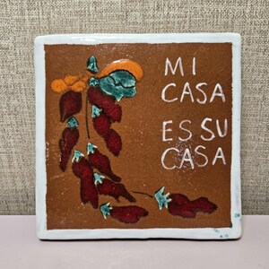 May include: Square ceramic tile with a brown background and white border. The tile features a stylized illustration of red chili peppers with green and orange accents. The text "MI CASA ES SU CASA" is written in white.