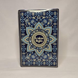 May include: A rectangular book with a dark blue cover featuring a complex, symmetrical mandala-style pattern in shades of light blue, cream, and navy. The center of the design displays the text Arabian Nights in a stylized, decorative font.
