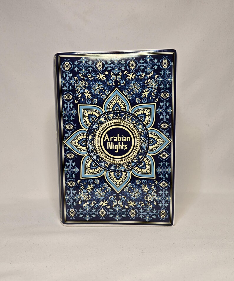 May include: A rectangular book with a dark blue cover featuring a complex, symmetrical mandala-style pattern in shades of light blue, cream, and navy. The center of the design displays the text Arabian Nights in a stylized, decorative font.