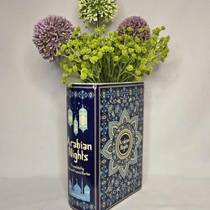 May include: A rectangular ceramic vase designed to resemble a book titled Arabian Nights, featuring dark blue surfaces with intricate gold and light blue patterns, hanging lantern illustrations, and a mosque silhouette, filled with purple and green flowers.