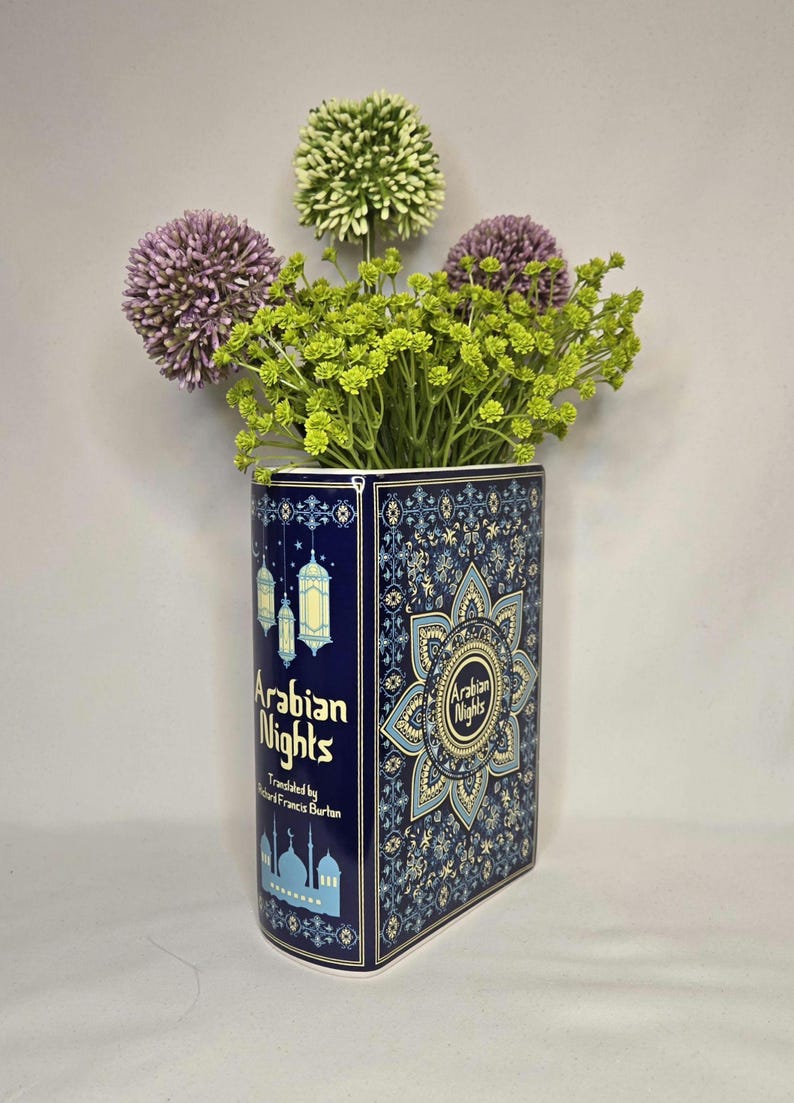 May include: A rectangular ceramic vase designed to resemble a book titled Arabian Nights, featuring dark blue surfaces with intricate gold and light blue patterns, hanging lantern illustrations, and a mosque silhouette, filled with purple and green flowers.
