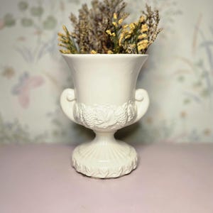 Puede incluir: Un jarrón de cerámica blanco con base de pedestal y dos asas. El jarrón presenta un relieve decorativo de uvas y hojas. El jarrón está lleno de flores secas y se encuentra sobre una superficie rosa.