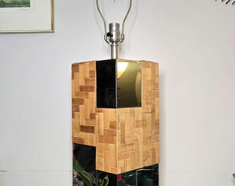 May include: A rectangular lamp with a woven bamboo base and mirrored accents. The lamp has a silver neck and a metal lamp holder. The base has a geometric design with mirrored squares. The lamp is on a wooden table.