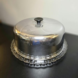 May include: A round cake dome with a polished silver metal cover featuring a small black knob handle on top. The cover sits securely on a clear glass plate that has a decorative scalloped edge design around the entire circular perimeter.