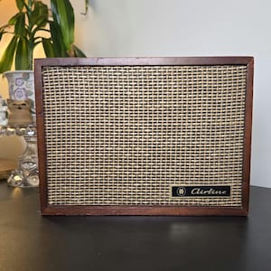 May include: A rectangular speaker with a dark brown wooden frame and a woven beige fabric grille. A small black rectangular badge with gold trim is located in the bottom right corner, featuring the word Airline in script and a small logo.
